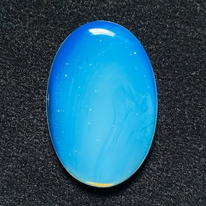 Peut inclure: Un cabochon de pierre précieuse de forme ovale avec une surface lisse et polie. La pierre présente une couleur bleue translucide et vibrante qui passe à une teinte plus claire et pâle vers le bord inférieur, avec de subtiles inclusions blanches.