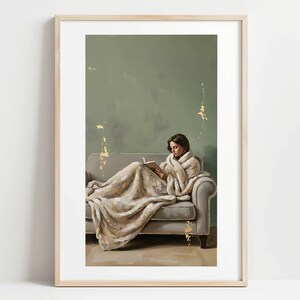 May include: A framed print depicts a person reading on a gray sofa, draped in a cream-colored blanket. The background is a muted green with gold accents. The artwork evokes a sense of comfort and relaxation.