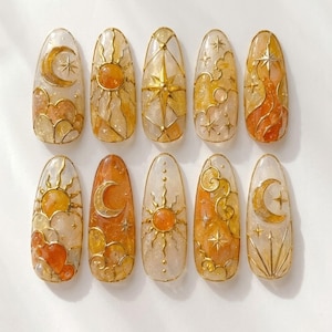 May include: A set of ten almond-shaped press-on nails featuring a celestial theme with gold metallic line art. The designs include suns, moons, stars, and clouds set against a translucent, glittery amber and cream-coloured resin base.