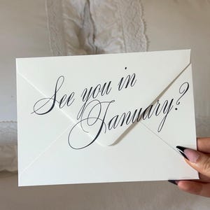 May include: A white rectangular envelope with a pointed flap featuring the black cursive text See you in January? printed across the center. The envelope is made of smooth paper and displays a formal script font style for the message.