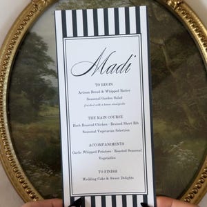 May include: A rectangular menu card with black and white vertical stripes at the top and bottom. The text reads Madi, TO BEGIN, Artisan Bread & Whipped Butter, Seasonal Garden Salad, THE MAIN COURSE, Herb Roasted Chicken, Braised Short Rib, Seasonal Vegetarian Selection, ACCOMPANIMENTS, Garlic Whipped Potatoes, Roasted Seasonal Vegetables, TO FINISH, Wedding Cake & Sweet Delights.