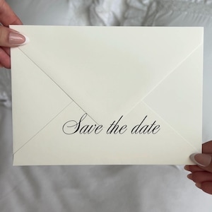 May include: A rectangular off-white envelope featuring a pointed flap and the text "Save the date" printed in a black, elegant, cursive script font centered on the lower portion of the envelope surface, showcasing a clean and classic design.