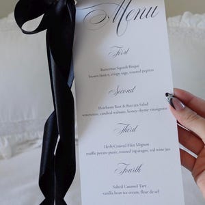 May include: A tall, rectangular white menu card with black text and a black satin ribbon tied through a hole at the top. The menu lists four courses: Butternut Squash Bisque, Heirloom Beet & Burrata Salad, Herb-Crusted Filet Mignon, and Salted Caramel Tart.