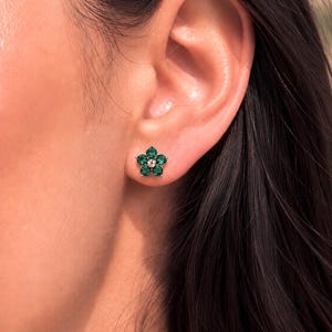 Può includere: Un orecchino a lobo a forma di fiore con cinque pietre rotonde sfaccettate di colore verde intenso, disposte come petali attorno a una piccola perla centrale color argento. L'orecchino è inserito in una montatura metallica sul lobo.