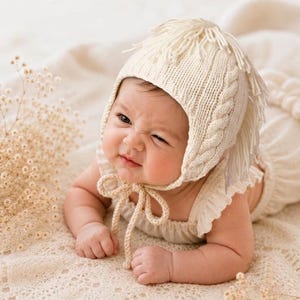 May include: A cream-colored knit bonnet featuring a textured cable knit pattern, a fringe detail along the top, and two long braided ties that fasten under the chin. The bonnet is crafted from a soft yarn and provides full head coverage.
