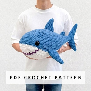 May include: A blue and white crocheted shark plush toy with black plastic eyes and a purple stitched mouth. The shark features a dorsal fin, pectoral fins, and a tail fin. The text PDF CROCHET PATTERN is displayed at the bottom of the image.