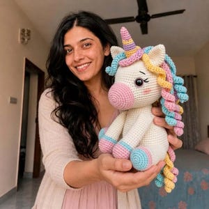 May include: A handmade crochet unicorn toy featuring a cream-colored body, a pink snout, and a spiraled horn in pastel yellow, pink, and blue. The unicorn has a multi-colored curly mane and tail in shades of blue, yellow, and pink, with black eyes.