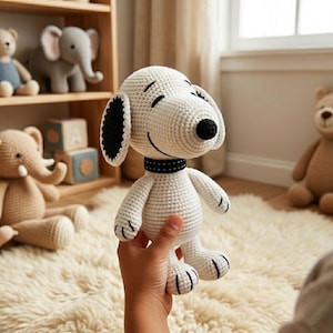 May include: A white crocheted Snoopy plush toy with black ears, nose, and collar. The smiling dog is held in a hand, with other stuffed animals and wooden blocks visible in the background. The toy is approximately 20 cm tall.