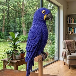 Puede incluir: Una escultura de loro azul hecha a mano, con pico negro y detalles amarillos, posada sobre un soporte de madera. El ave está hecha de hilo texturizado. La escultura se exhibe en interiores, una pieza decorativa cerca de una ventana.
