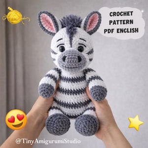 May include: A handmade crochet zebra plushie with black and white stripes, gray accents, and a gray mane. The text "CROCHET PATTERN PDF ENGLISH" is in a speech bubble. The plushie is held in hands. The shop's name is @TinyAmigurumiStudio.