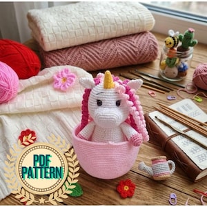 Pode incluir: Unicórnio de crochê com crina rosa e chifre amarelo em uma cesta rosa. A imagem inclui fios, agulhas de crochê e um pequeno regador de crochê. O texto "PDF PATTERN" é visível em um emblema circular.