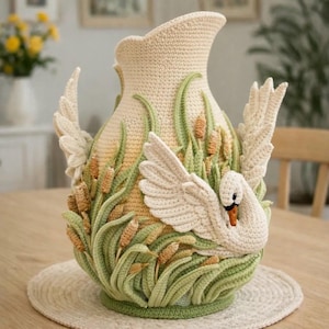 May include: A handcrafted, cream-colored crochet vase featuring a swan design. The vase has a wide base and a flared top. The swan is depicted with outstretched wings, surrounded by green and brown cattails and reeds. The vase is approximately 12 inches tall.