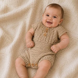 May include: A beige crochet romper featuring short sleeves, a round neckline, and a button-down placket on the chest. The garment includes three small wooden buttons at the bottom crotch area for closure, showcasing a textured, hand-stitched pattern.