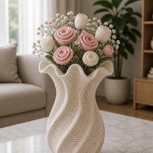 Op de afbeelding: Een crèmekleurige, gehaakte vaas met een gerimpelde bovenkant bevat een boeket handgemaakte bloemen. Het boeket bevat roze en witte rozen, witte tulpen en kleine witte vulbloemen. De vaas staat op een tafel met marmeren blad.