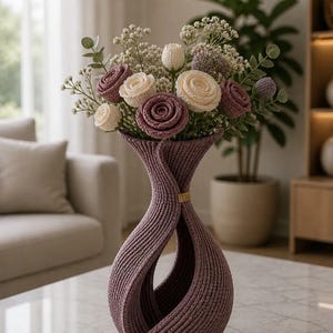 May include: A decorative purple vase with a unique twisted design, holding a bouquet of crocheted flowers in shades of white, cream, and purple. The vase has a gold band near the top and base, and is sitting on a marble table.