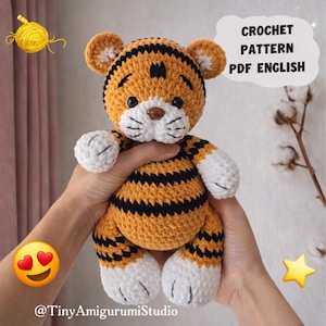 Puede incluir: Un tigre amigurumi tejido a crochet en naranja, negro y blanco. El tigre tiene rayas negras, patas blancas y un hocico blanco. El texto "CROCHET PATTERN PDF ENGLISH" está en una burbuja. El identificador de Instagram @TinyAmigurumiStudio está en la parte inferior.
