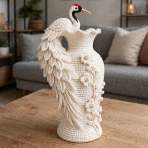 Könnte beinhalten: Handgefertigte, cremefarbene Häkelvase mit Kranich-Design. Der Kranich hat einen roten Kopf, schwarz-weiße Markierungen und einen beigen Schnabel. Die Vase ist mit floralen Verzierungen und einem gerüschten Oberteil versehen, was sie zu einem einzigartigen Dekorationsstück macht.