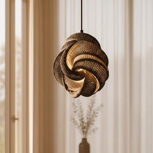 May include: A modern, decorative pendant light fixture with a unique, swirling design. The light is made of a dark, textured material with a perforated pattern, and it hangs from a black cord. The interior glows with warm light, creating a soft ambiance.