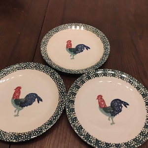May include: Three white ceramic plates with a green dotted border. Each plate features a rooster illustration with a red comb, blue feathers, and green body. The plates are arranged on a dark brown wooden surface.
