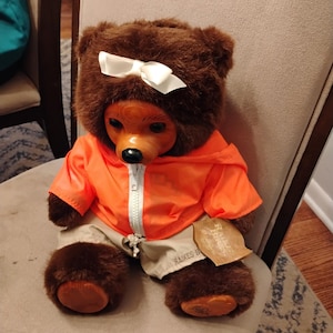 Op de afbeelding: Een bruine teddybeer met een witte strik, gekleed in een oranje jas en kaki broek. De beer heeft een houten gezicht en zit op een stoel. Een label is aan het been van de beer bevestigd.