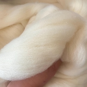 Op de afbeelding: Een grote, zachte massa ongesponnen, gebroken witte wolvacht. De vezels zijn dicht op elkaar gepakt en hebben een pluizige, wolkachtige textuur met een consistente crèmekleur door het hele bundel, wat de natuurlijke krul van het materiaal toont.