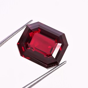 May include: A faceted, rectangular-cut, deep red gemstone held by silver tweezers. The gem has a vibrant, polished surface and a clear, internal structure, reflecting light. The gem is a rich, saturated color, with sharp, defined edges.