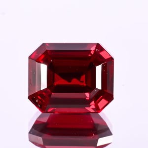 Può includere: Una gemma rosso scuro tagliata a forma rettangolare. La gemma ha una superficie brillante e riflettente, su sfondo bianco. Le sfaccettature creano un gioco di luci e ombre, esaltando il suo colore intenso.