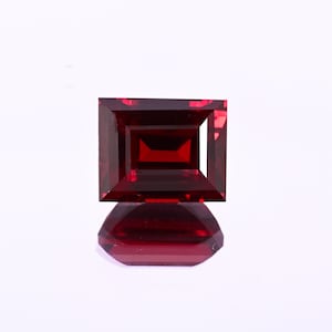 May include: A faceted, rectangular-cut, deep red gemstone with a smaller rectangular facet inside. The gemstone is displayed on a reflective surface, creating a mirror image. The gem is a rich, dark red colour.