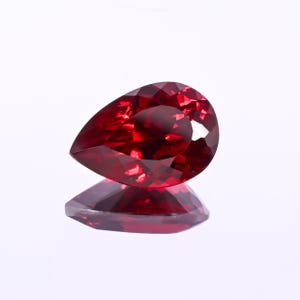 Può includere: Una gemma sfaccettata, a forma di pera, di colore rosso intenso. La gemma è molto riflettente, con molteplici sfaccettature che catturano la luce. La pietra è posta su uno sfondo bianco, con il suo riflesso visibile sotto.
