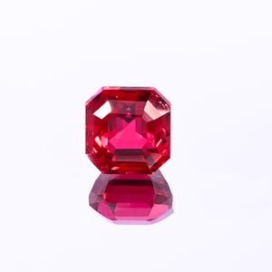 May include: A faceted, rectangular-cut, red gemstone with a brilliant shine. The gem is set against a white background, with its reflection visible below. The gemstone's facets catch the light, creating a sparkling effect.