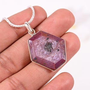 Può includere: Una collana d'argento con un pendente con una grande pietra preziosa sfaccettata, di colore rosso-viola. La gemma ha una forma geometrica unica e un motivo interno visibile. Il pendente è incastonato in una lunetta d'argento e appeso a una sottile catena d'argento.