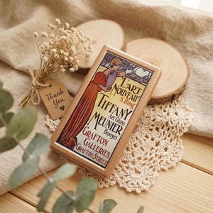 May include: A rectangular metal case featuring a vintage-style graphic print of a person in a long red dress. The text reads L'ART NOUVEAU S. BING TIFFANY MEUNIER ART GLASS BRONZES GRAFTON GALLERIES GRAFTON STREET. The case has a metallic gold finish.