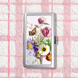 May include: A rectangular silver-toned metal case featuring a printed illustration of a bouquet of flowers in shades of purple, white, pink, and red, accompanied by a brown and orange butterfly, all set against a white background.
