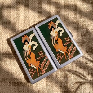 May include: Two rectangular silver-toned cases featuring a vintage-style graphic print of a woman in an orange dress with white ruffles. The text reads: Chas. H. Yale's Everlasting 20th Edition and Best Ever The Devil's Auction in stylized lettering.