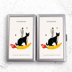 May include: Two rectangular silver metal cases featuring a graphic print of an astronaut standing on a black and white cat, which is riding a yellow banana with flames at the end. The text reads DAYDREAMER and Turning your imagination into reality.