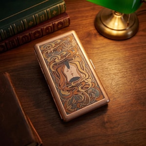 May include: A rectangular metal cigarette case featuring an intricate Art Nouveau style illustration of two faces in profile kissing, surrounded by flowing golden hair and floral motifs in shades of amber, cream, and muted blue on a copper frame.