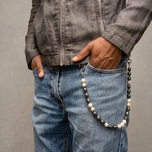 Puede incluir: Una cadena para cartera con cuentas alternas de color gris oscuro y blanco, sujeta a la trabilla del cinturón y al bolsillo de unos vaqueros azules. La cadena está formada por una serie de cuentas lisas y redondas, aseguradas con cierres metálicos plateados.