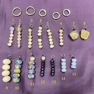 May include: A collection of fifteen jewelry charms and five silver-tone jump rings arranged in rows. The charms feature various beads including white pearls, yellow stones, multicolored floral patterns, dark purple, and light blue cylindrical beads.