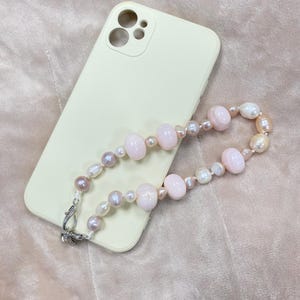 May include: A pale yellow smartphone case with a camera cutout, paired with a beaded phone charm featuring a mix of round, smooth, light pink beads and irregular, lustrous white and soft lavender pearls, finished with a silver metal lobster clasp.