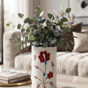 Könnte beinhalten: Eine zylindrische Vase aus weißem Marmor mit einem eingelegten Blumenmuster aus roten und blauen Steinakzenten. Die Vase ist mit grünen Eukalyptuszweigen gefüllt und steht aufrecht, wobei sie eine glatte, polierte Oberfläche und einen flachen Boden zeigt.