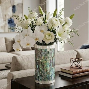 Könnte beinhalten: Eine zylindrische Vase mit einem schillernden Abalone-Muschel-Mosaik-Finish enthält ein großes Arrangement aus weißen Blumen, darunter Orchideen, Lilien und Ranunkeln. Die Vase hat einen glatten weißen Rand an der Ober- und Unterkante.