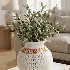 Könnte beinhalten: Eine weiße, kugelförmige Vase mit einem komplexen Gittermuster im unteren Bereich und einem farbenfrohen Blumen-Inlay am Hals. Die Vase ist mit einem üppigen Arrangement aus grünen Olivenzweigen mit kleinen, dunklen Oliven gefüllt.