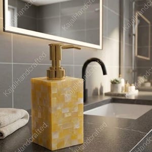 Könnte beinhalten: Dekorativer Seifenspender mit goldener Pumpe und gelb-weißem Mosaikfliesen-Design. Der rechteckige Spender steht auf einer dunklen Arbeitsplatte in einem Badezimmer. Im Hintergrund sind Spiegel und Waschbecken zu sehen.