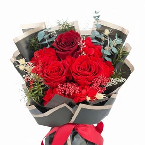 Puede incluir: Un ramo de rosas rojas y otras flores rojas envuelto en papel negro con un borde dorado. El ramo est&aacute; atado con una cinta roja. El arreglo incluye eucalipto y otras plantas verdes.