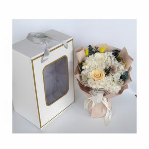 Puede incluir: Una caja de regalo blanca con un borde dorado y una ventana transparente, junto a un ramo de flores. El ramo presenta rosas blancas, crema y melocot&oacute;n y otras flores, envueltas en papel marr&oacute;n claro y atadas con una cinta.