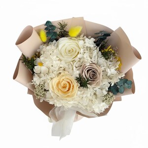 Puede incluir: Un ramo de flores preservadas envuelto en papel beige. El arreglo presenta hortensias blancas, rosas crema y beige, y ramitas de vegetaci&oacute;n y acentos amarillos. Una cinta blanca asegura la base del ramo.