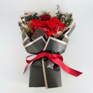 Puede incluir: Un ramo de rosas rojas y vegetaci&oacute;n, envuelto en papel negro con ribetes dorados. Una cinta roja ata la base. El arreglo presenta vibrantes rosas rojas y varios elementos verdes y secos, creando un contraste llamativo.