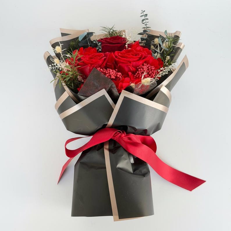 Puede incluir: Un ramo de rosas rojas y vegetaci&oacute;n, envuelto en papel negro con ribetes dorados. Una cinta roja ata la base. El arreglo presenta vibrantes rosas rojas y varios elementos verdes y secos, creando un contraste llamativo.