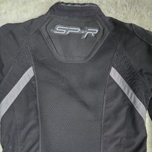 Peut inclure: Veste de moto noire avec des d&eacute;tails gris et les lettres "SP-R" au dos. La veste pr&eacute;sente un panneau en mesh et des bandes grises r&eacute;fl&eacute;chissantes sur les manches. Le col est montant.