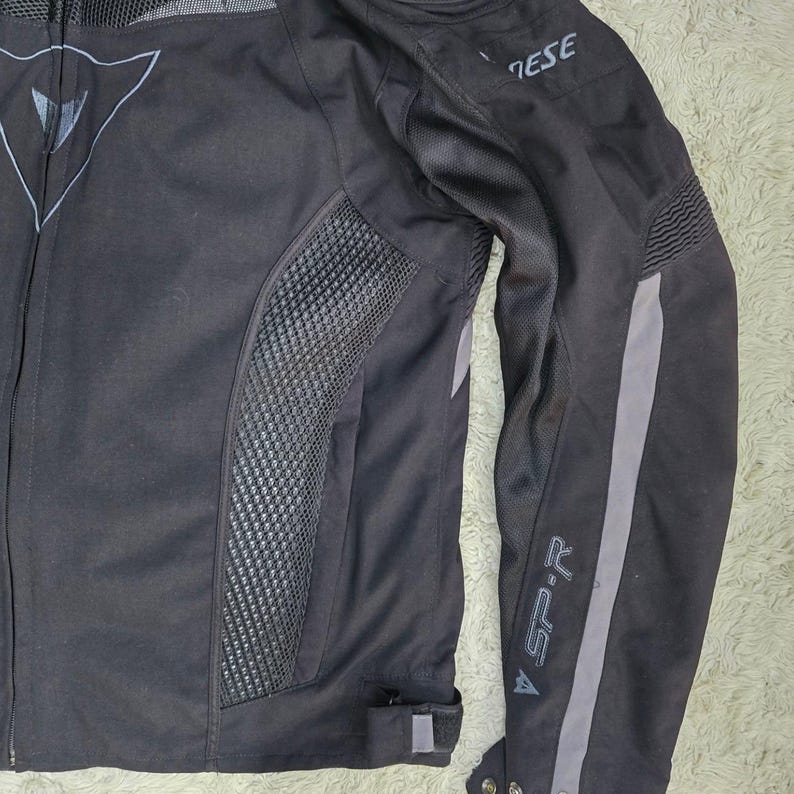 Peut inclure: Veste de moto noire avec des d&eacute;tails gris et des panneaux en mesh. Comprend le logo de la marque sur la poitrine et la manche. La veste a une fermeture &eacute;clair et une coupe ajust&eacute;e, pour la moto.